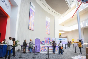 A well-lit convention center lobby with people registering at the "AHIP 2023" desk. Large, colorful banners hang on the walls. Casual, busy atmosphere.
