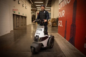 A security officer rides a three-wheeled Segway in a building corridor. He wears a helmet and uniform. A red wall displays text and building silhouettes.