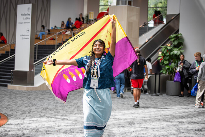 A smiling young person confidently walks through a busy lobby, wearing a blue Indigenous ribbon shirt and skirt, holding up the yellow and purple flag of the Tohono O'odham Nation.