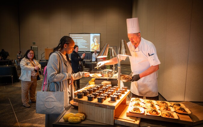 A chef behind a serving table full of food as an event attendee peruses the options.