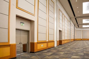 Spacious, empty ballroom with high ceilings, patterned carpet, large panel-covered walls, and double exit doors. Bright and formal atmosphere.