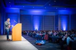 A speaker in a gray jacket addresses a large, attentive audience from a podium under blue lighting. The room is spacious with an engaged crowd seated at tables.