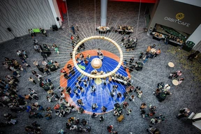 A 3-foot polished brass pendulum swings above a 15-foot gilded ring featuring tapered rods that extend both outward and inward. Below, a gathering of event guests mingles over a 30-foot circular blue terrazzo floor inlaid with brass rings and colored stone planets representing a fantasy solar system.