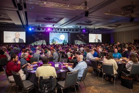 A large ballroom filled with attendees seated at round tables, facing a stage with a nature-themed backdrop. Two screens display a speaker.