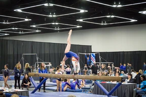 Gymnast performs a handstand on balance beam in a busy indoor arena. Judges observe intently, while other gymnasts and spectators watch. Energetic mood.