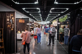 People explore a bustling craft fair with various stalls displaying pottery and art. The scene is lively, with bright overhead lights and colorful decor.