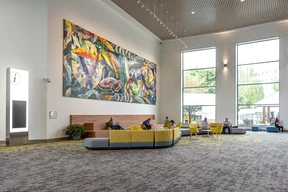 Spacious lobby with a vibrant, abstract mural on the wall. People sit on modern yellow and gray couches under large windows, creating a relaxed atmosphere.