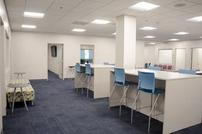 Modern space with blue carpet and white walls. High tables with blue chairs are centered. Benches with patterned cushions line the wall, emitting a bright, organized feel.
