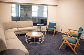 Comfortable lobby with a gray sectional, round coffee table, and two blue armchairs. Light filters through large windows, creating a calm, inviting atmosphere.