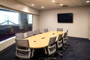 A modern conference room with an oval wooden table and eight gray ergonomic chairs. A wall-mounted TV and large windows give a professional, open feel.