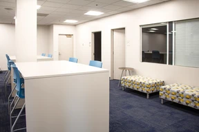 Modern office space with white walls, high tables, blue chairs, and geometric-patterned benches. Soft lighting and a calm, clean atmosphere.