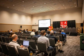 Conference room with attendees seated facing a stage. A speaker presents at a podium with "IAVM" on banners. Attentive audience, one person using a laptop.