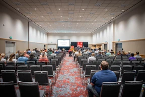 A large conference room with rows of chairs filled with attendees facing a presentation screen and a stage with speakers. Calm, focused atmosphere.