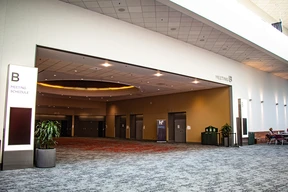 Spacious convention center hallway with beige and red carpet, glass doors, and a hanging light. Sign on the left reads "Meeting Schedule B."