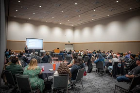 A conference room filled with seated attendees facing a presenter and a projection screen. The atmosphere is attentive and focused.