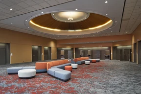 Spacious lobby with modern gray and orange modular seating under a large circular light fixture. Red and gray carpet, beige walls, and labeled doors.