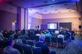 Audience attentively listens to a speaker in a dimly lit conference room. A presentation is projected on a screen at the front, creating an engaging atmosphere.