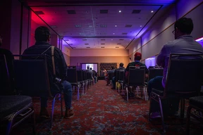 Audience in a dimly lit conference room with purple and red lighting, attentively facing a stage. The mood is focused and professional.