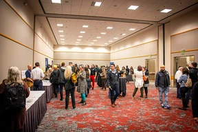 A large conference room filled with people engaged in conversation. Groups are gathered near tables with white tablecloths. The atmosphere is lively and social.