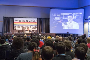 Audience faces a stage with a speaker at a table and a large screen displaying a video. Room features retro gaming theme. Engaged and attentive atmosphere.