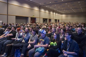 A large, cheerful audience at a convention. Attendees of various ages sit closely packed, smiling and laughing, creating a lively and engaging atmosphere.
