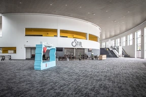 Spacious modern lobby with gray carpet, curved white walls, and large windows. "Welcome" sign and "Stir" cafe create a welcoming atmosphere.