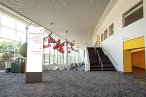 Spacious lobby with a curved ceiling and long staircase. A white sign lists areas and amenities. Bright red art installation contrasts with gray decor.