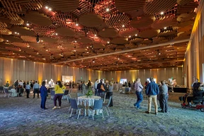 Spacious event ballroom with a geometric wooden ceiling and warm lighting. Groups of people mingle around tables, creating a lively and inviting atmosphere.