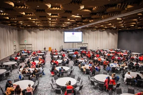Spacious ballroom with round tables, attendees mostly in red shirts, facing a presentation screen. Mood is focused and collaborative. Ceiling features geometric patterns.