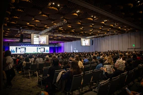 A large audience seated in a ballroom watches presentations on multiple screens. The atmosphere is focused and engaged, with a modern ceiling design.