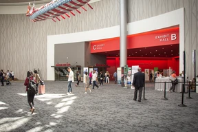 Spacious convention center entrance with "Exhibit Hall B" and "C" in bold red signs. People walk through, suggesting a bustling event atmosphere.